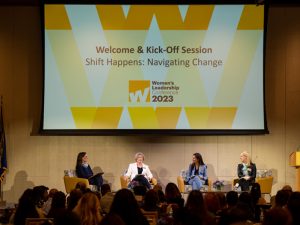 Four women are seated on chairs on a stage. The screen overhead reads: Welcome and Kick-Off Session, Shift Happens: Navigating Change, Women's Leadership Conference 2023. The sign on the podium reads: Adelphi University.