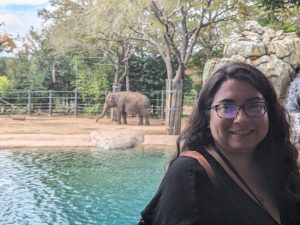 A woman wearing glasses in the foreground in front of an elephant in a zoo.