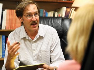 A man seated in front of a bookshelf is talking animatedly.
