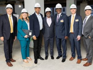 Group photo of seven people, six men and one woman, posed together wearing hard hats and formal business attire in a new construction space