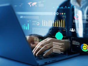 A person typing on a laptop with transparent digital dashboards overlaid in front of the screen, displaying charts, graphs, and analytics data.