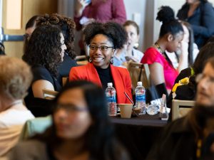 Natalie Ward, wearing a red blazer, sits at a table at the conferences while talking with another student. The two are surrounded other students at tables.