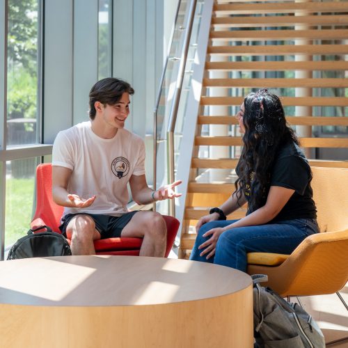 A group of five Adelphi students smiling in front of a university building holding laptops and books