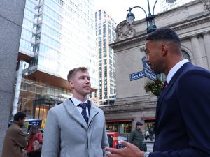 Two men in professional attire stand on a busy street corner in New York City, engaged in conversation. 