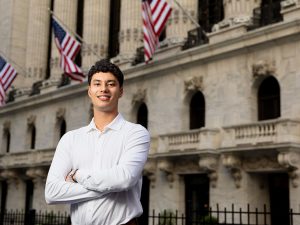 A male student in a collared shirt standing in a city setting with his arms crossed. 