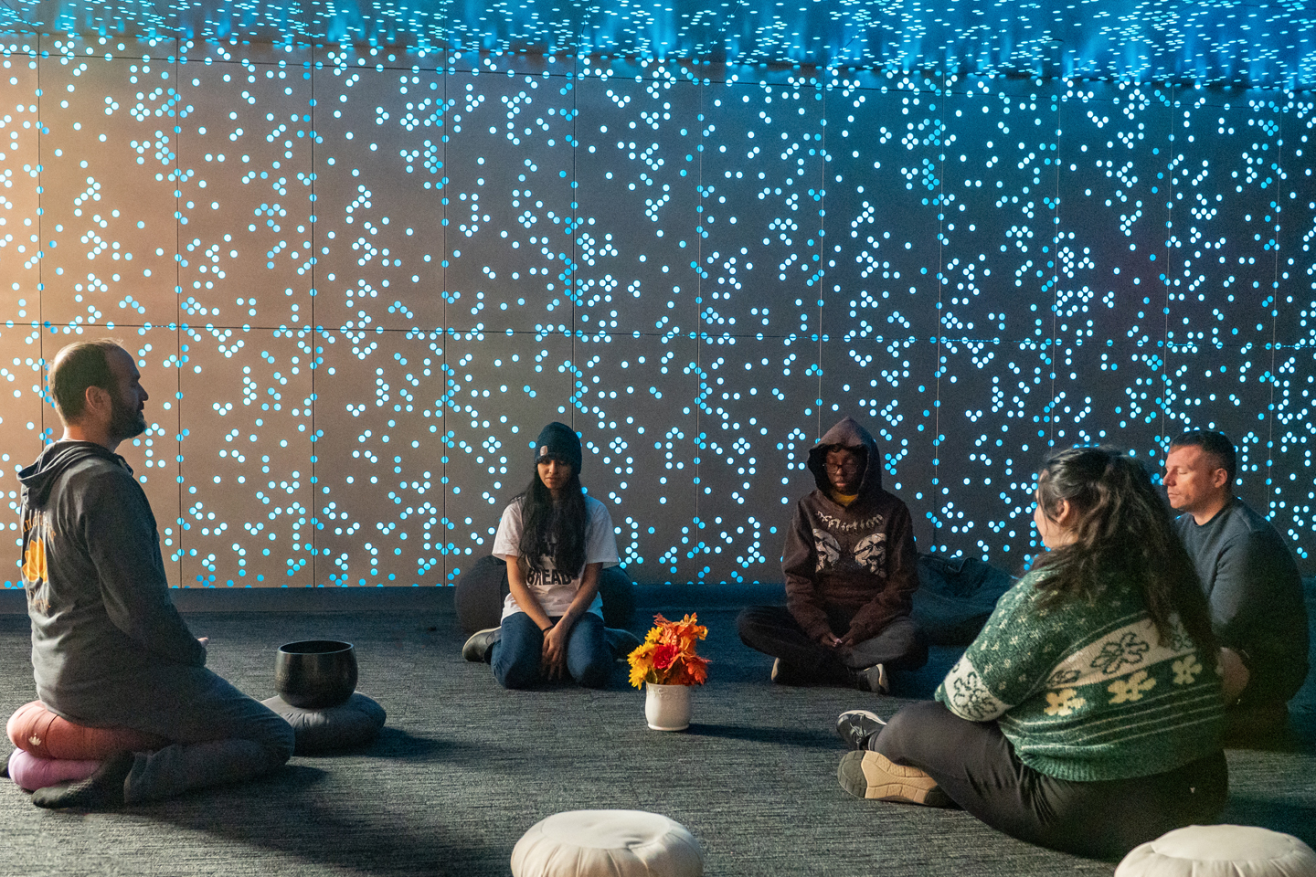 Four students and an instructor sit on the floor of the dimly lit Mindfulness Center. Quietly sparkling blue lights on the wall enhance the mood.