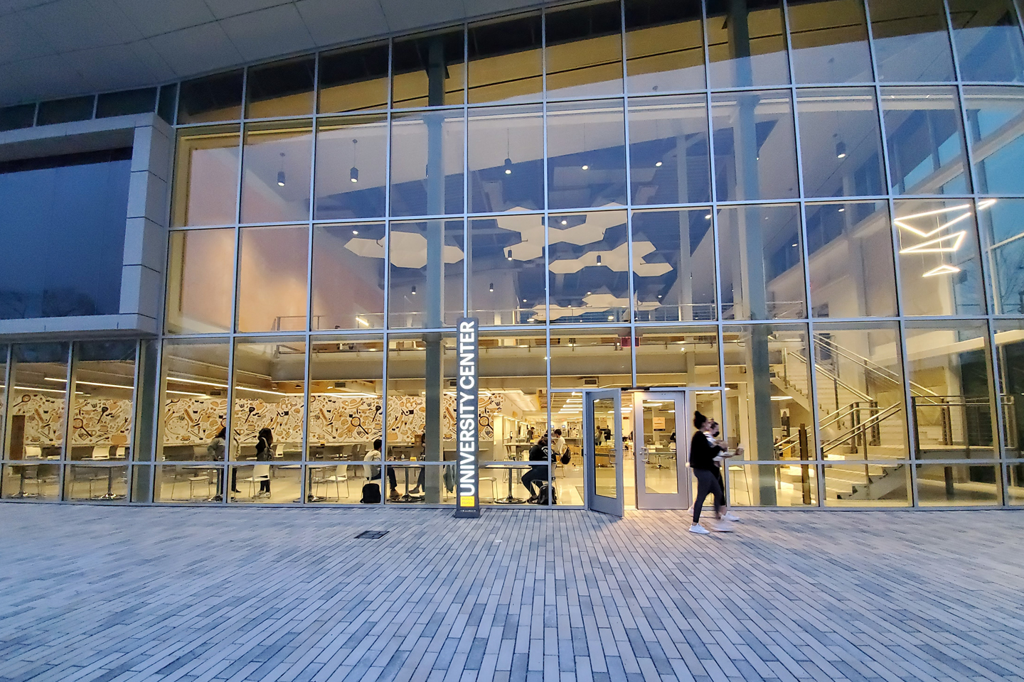 A student walks at dusk in front large windows University Center, which provide a view into the building's well-lit interior.