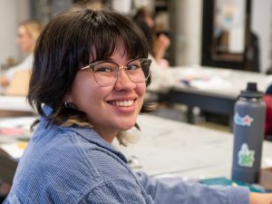 A young student smiles as she turns from a classroom desk to fact the camera.