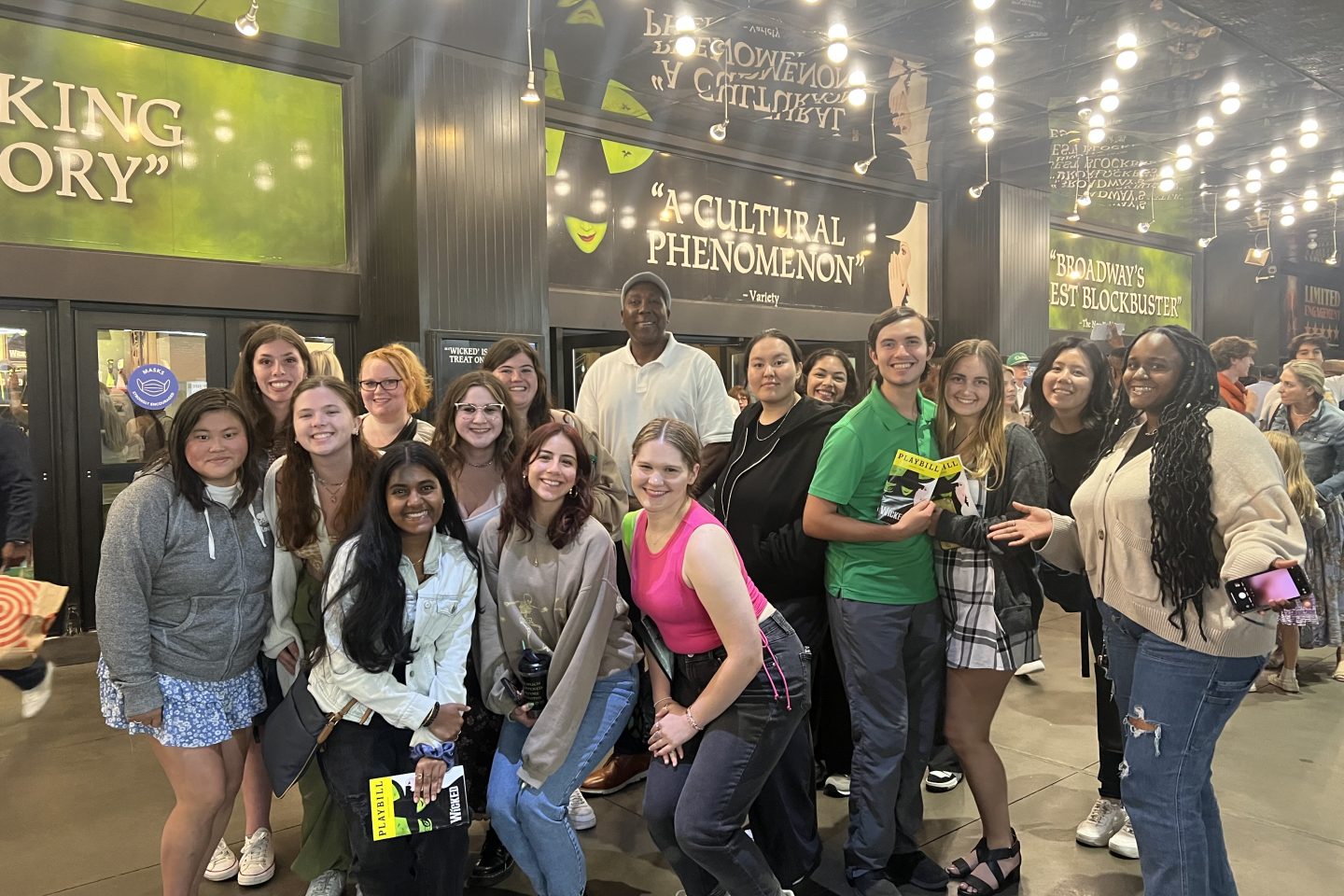 A group of eighteen people, predominantly younger individuals, standing close together and smiling at the camera are shown in an outdoor space beside a Broadway theater. Overhead are bright lights, and there is green signage in the background with large white text with the phrases "CULTURAL PHENOMENON," "WICKED STORY," and "BROADWAY'S BEST BLOCKBUSTER.
