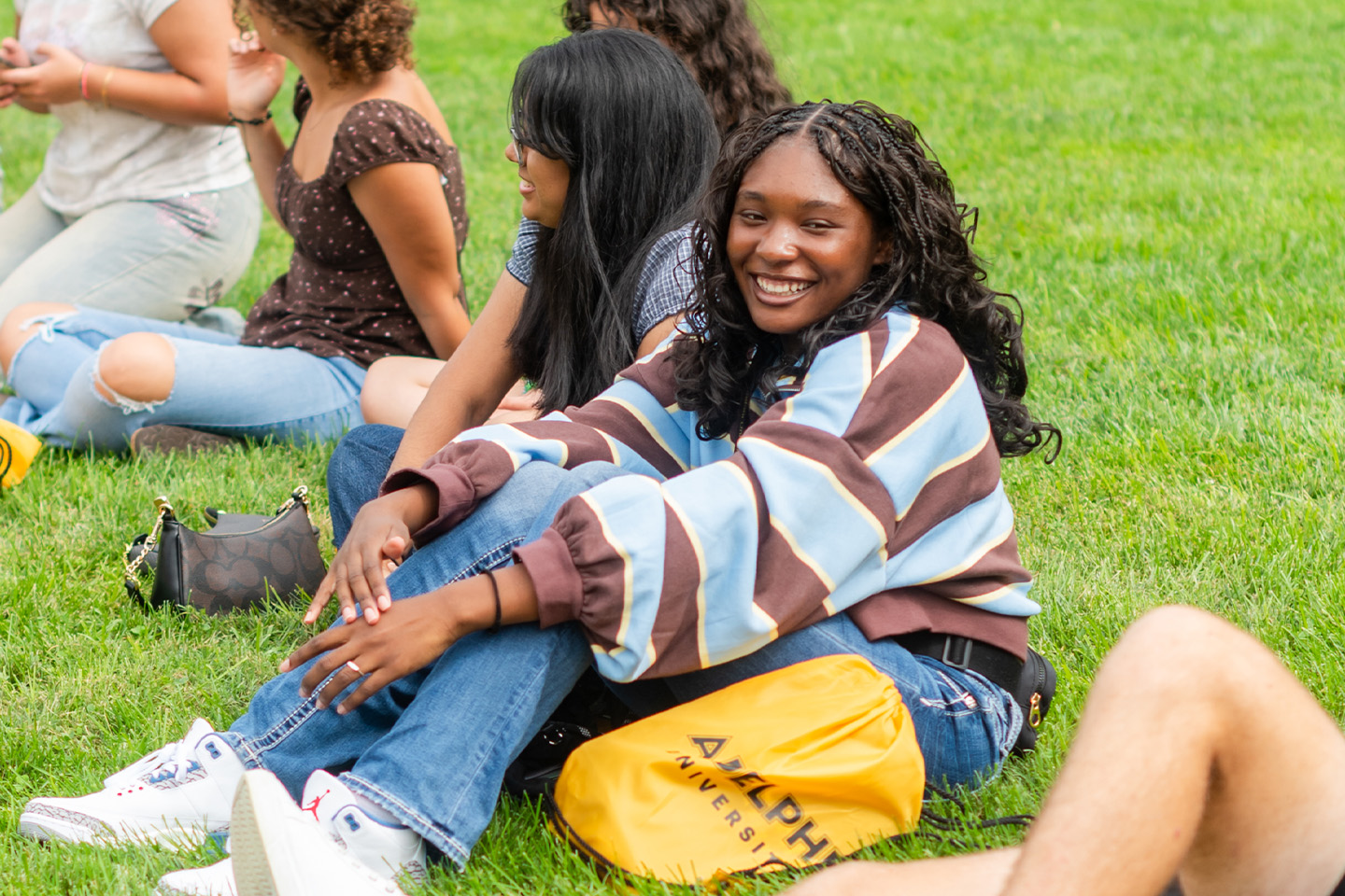 A handful of students sit on the lawn on a sunny day. One student smiles at the camera.