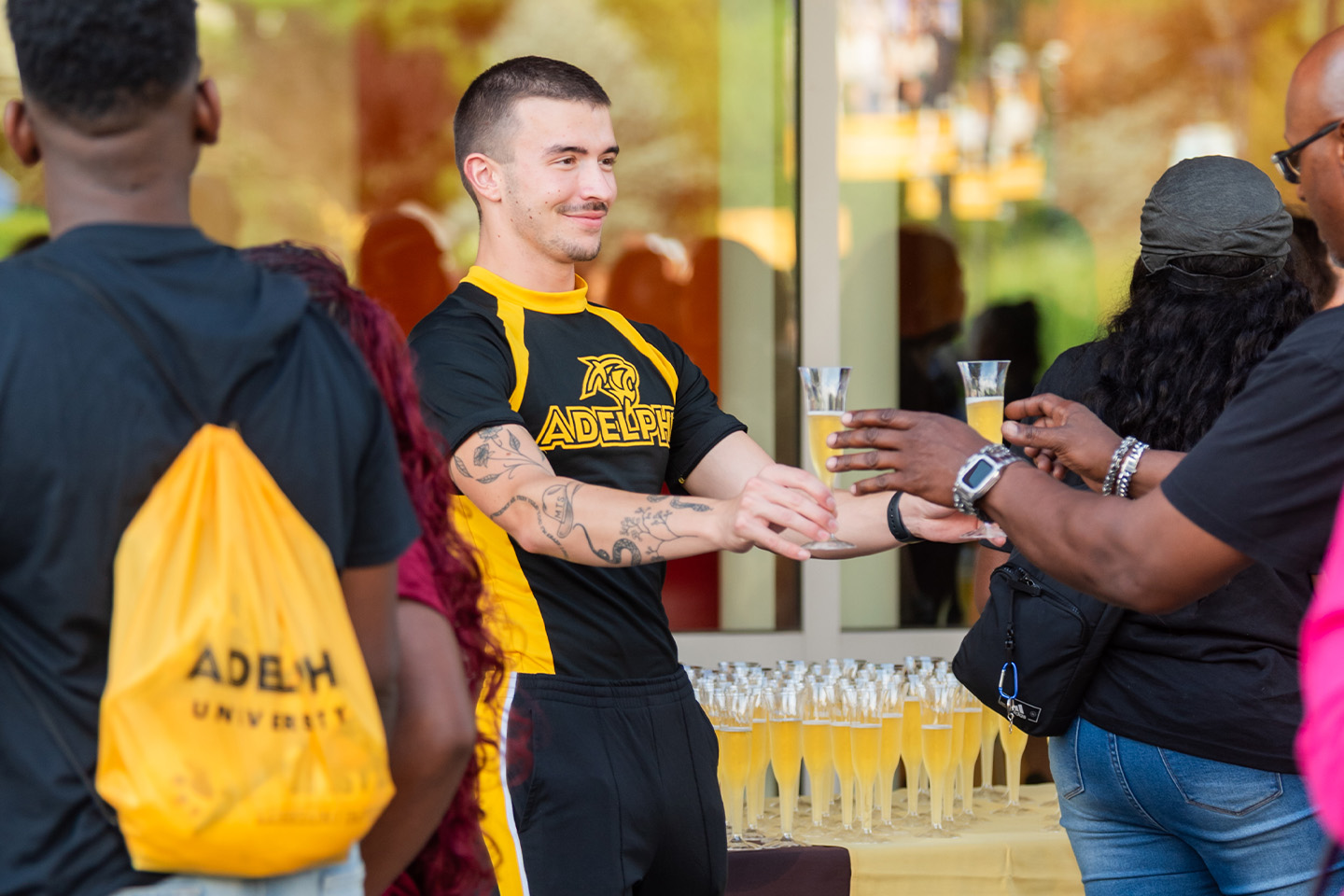 Braden, wearing a black and gold Adelphi shirt, hands two cups of cider to a parent.