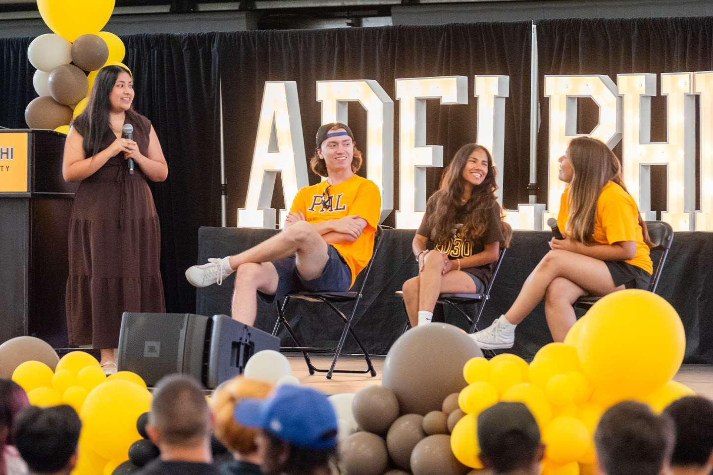 Three students sit on stage with an Adelphi banner behind them. The session's host stands at their right while holding a microphone.