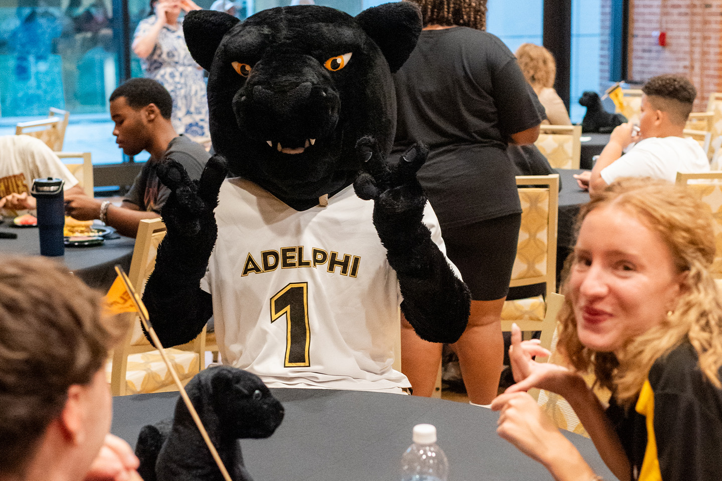 The Adelphi mascot, a black Panther in an Adelphi jersey with a No. 1 on it, sits at a table with two new students.