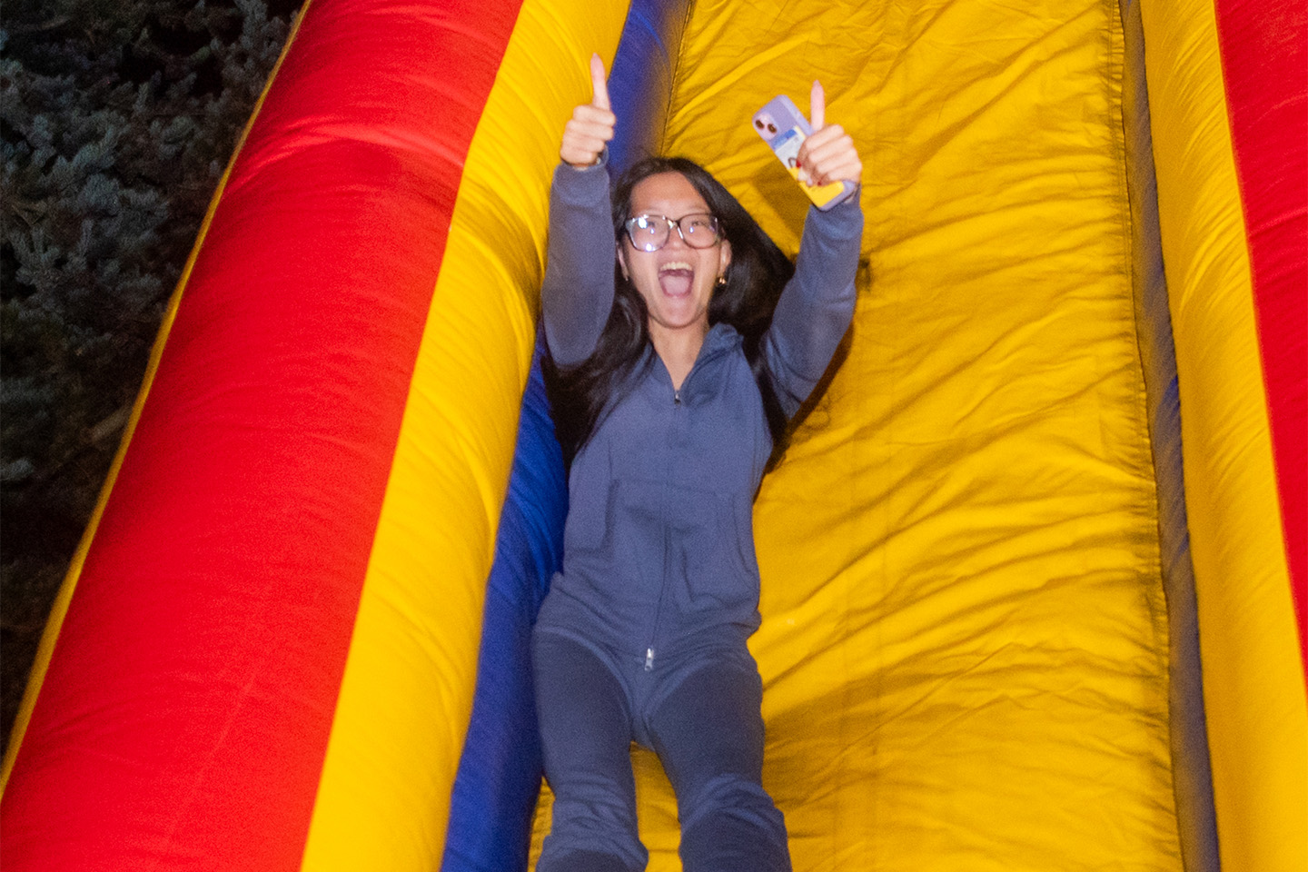 A student whisks down an inflatable slide with her arms overhead and flashing a thumbs-up.