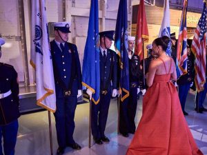 A row of military servicemen in full-dress uniform holding flags at a gala, greeting a woman wearing a formal, full-length red gown, whose back is to the camera