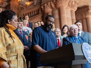 Walters stands at the microphone on a pedestal fronted by the seal of the State of New York. He is surrounded by veterans, many wearing American Legion caps. The scene is inside the ornate lobby of the historic state capitol.  