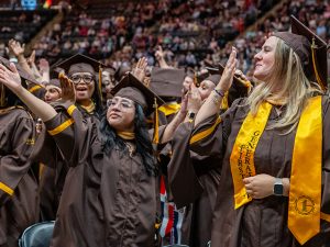 In a crowd of graduates on Commencement day, one grad blows a kiss and the other waves to family and friends.