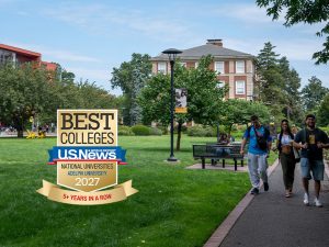 A group of students walking along a path with campus buildings and landscape in the background, including an overlay of a US News & World Report Best Colleges badge in the bottom left corner.