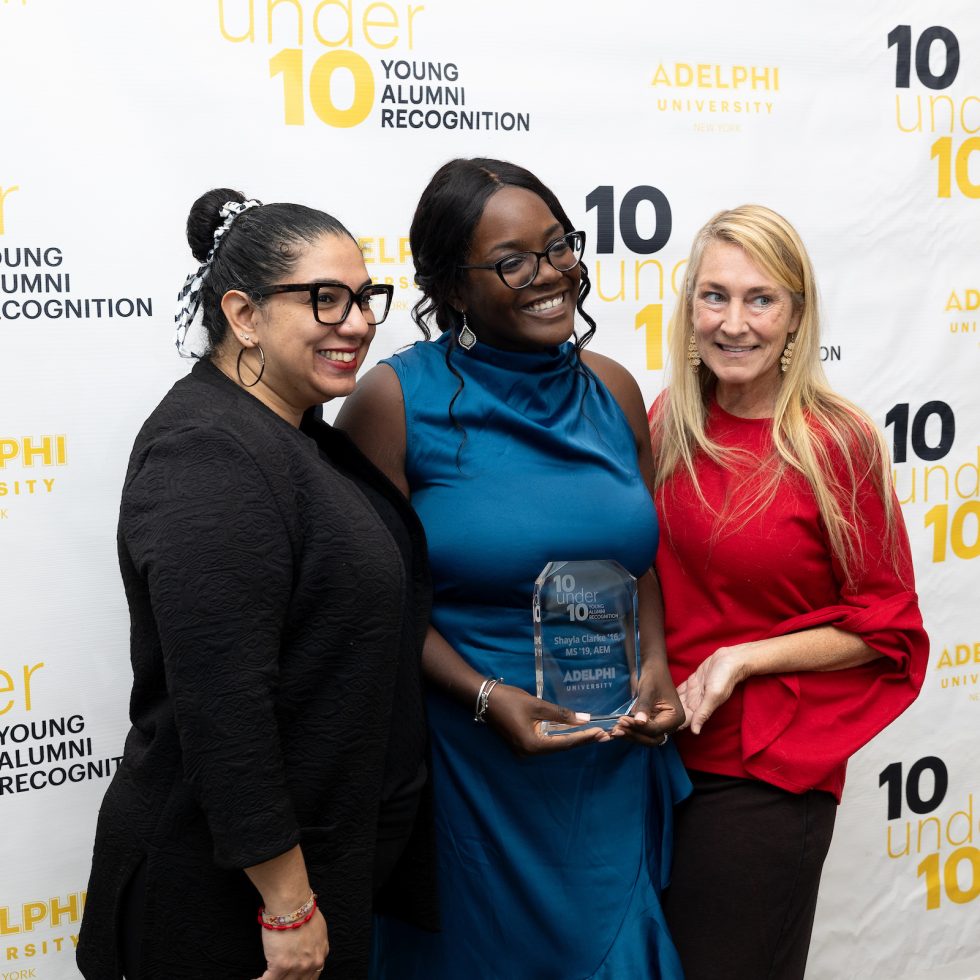 Adelphi 10 under 10 recipient Shayla Clarke ‘16, MS ‘19 (center) posing in front of step and repeat during award ceremony.