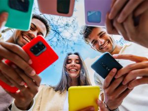 A group of friends stand in a circle holding colorful smartphones above their heads, photographed from below looking up. They smile down at the camera with blue sky and bare tree branches visible in the background.