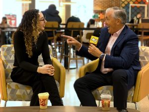 A young student, seated on the left, chats with a man in a suit on the right. They are drinking coffee and laughing casually. 