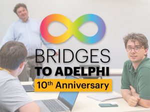 A Bridges student in an Adelphi physics class faces the camera. Two other students are seated across the table from him. A professor stands at the front of the class.