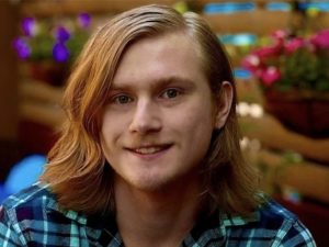 A young man with long auburn hair smiles at camera.