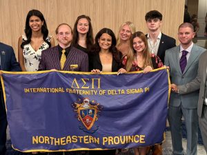A group of ten smiling young professionals stands together indoors, holding a large purple banner for Delta Sigma Pi, a professional business fraternity.