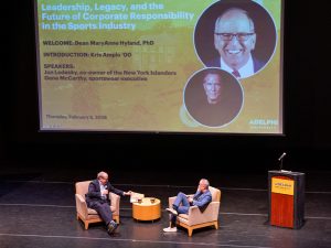 Two men sit in armchairs on a stage, talking, with a low table and coffee cups between them. Behind them, a large screen displays the event title "Leadership, Legacy, and the Future of Corporate Responsibility in the Sports Industry," hosted by Adelphi University's Distinguished Lecture Series, dated Thursday, February 5, 2026, alongside photos of the two speakers. A podium with a microphone stands to the right, and branded pull-up banners reading "Distinguished Lecture Series" flank the stage on both sides.