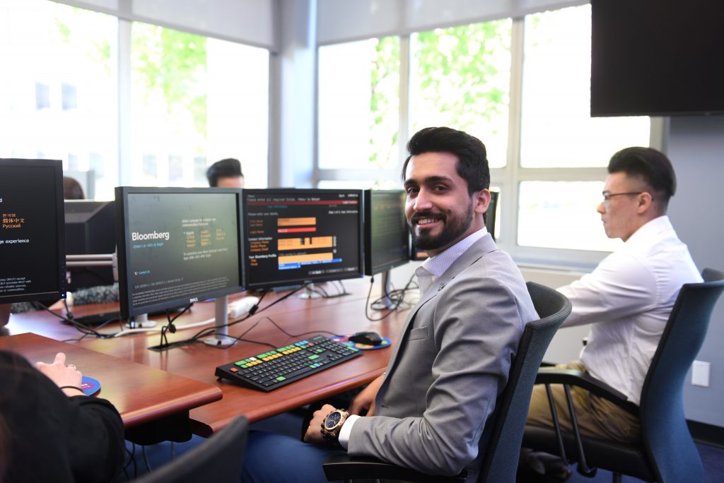 A smiling business student dressed in a light grey suit seated at a Bloomberg Terminal in the Adelphi University Trading Room in Hagedorn Hall.