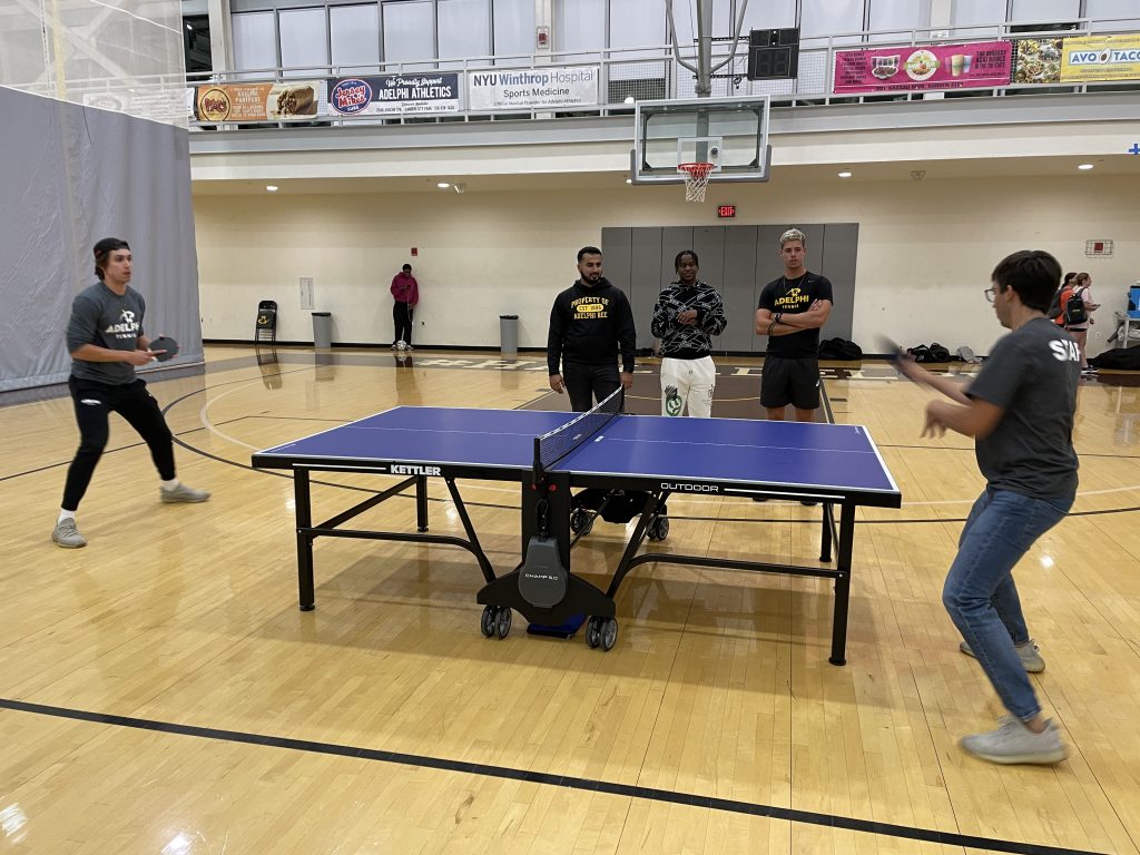 2 male students playing table tennis in the gym and 3 male students watching them play. 
