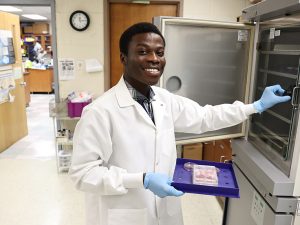 Samuel Sey in a well-lit lab. Wearing a lab coat and blue surgical gloves, he holds a tray with specimens on it, which he prepares to place in a refrigerated cabinet. He is looking at the camera and smiling.