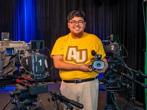 A young man in glasses and a gold shirt that reads AU—for Adelphi University— is smiling, holding a camera. On the left is a camera labeled 1 Autoscript, and on the right, a camera labeled 2.