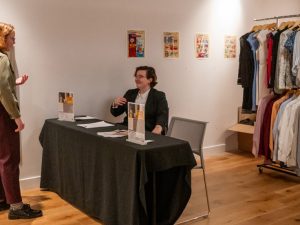 Woman greeting someone in a suit jacket who's sitting at a table with a clothes rack behind them