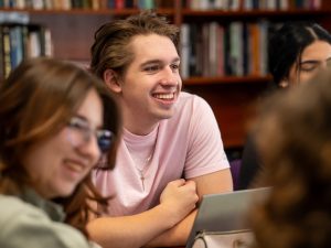 A student at a seminar table, with a student on each side of him in soft focus. Bookcases line the seminar room's walls.