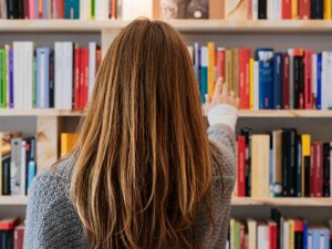 A woman with long brown hair, seen from behind, is reaching out toward a bookshelf filled with colorful books. She is wearing a gray sweater and appears to be selecting a book in a library or bookstore setting.