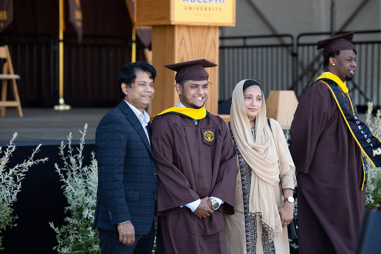 Graduating student poses with proud parents in front of commencement stage.