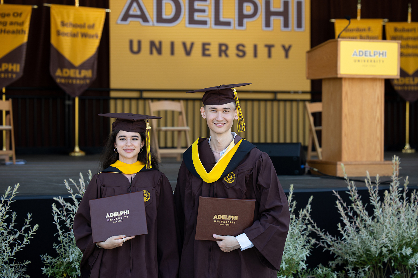 Two Adelphi graduates holding diplomas pose for a picture.