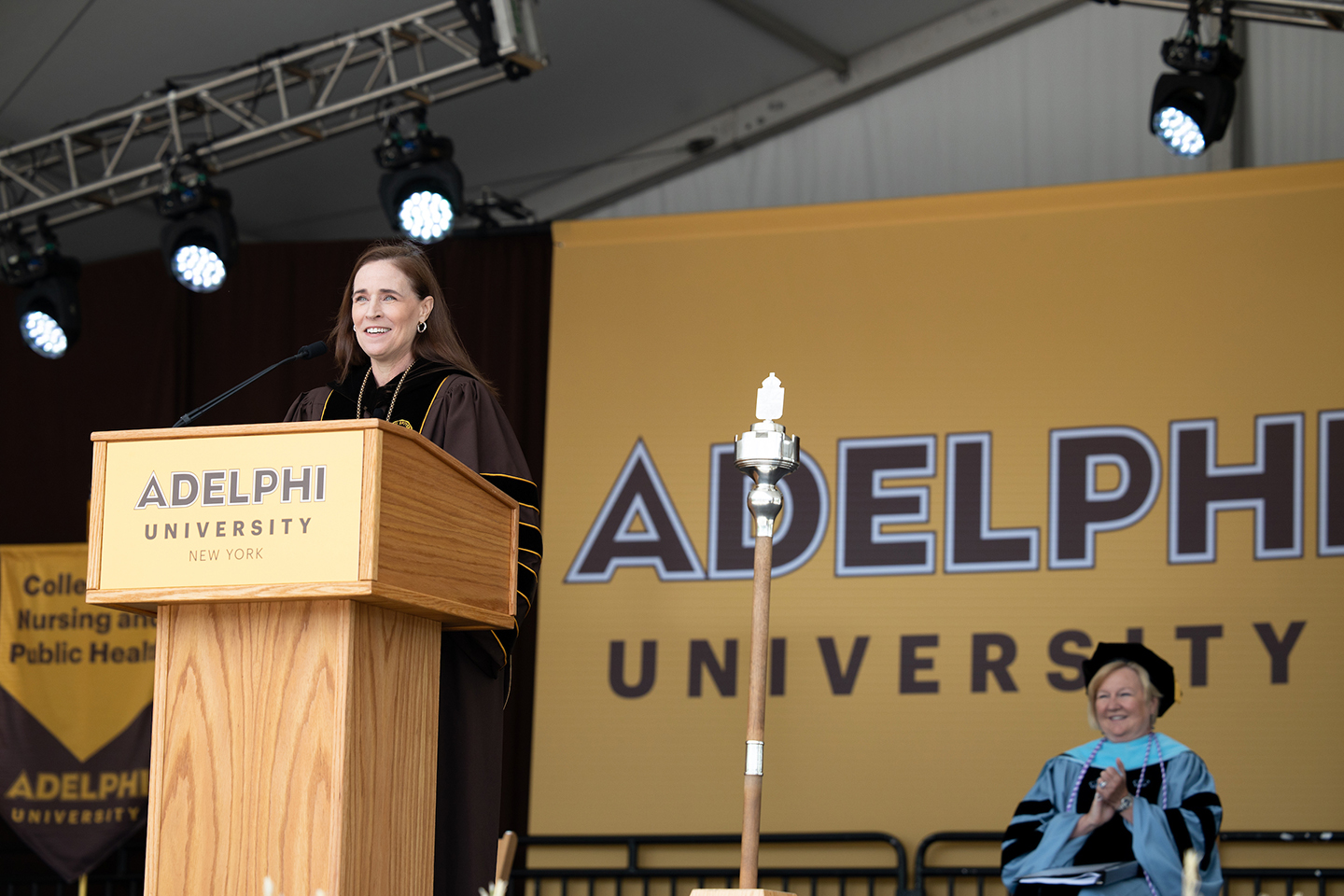 President Riordan standing behind a podium in a commencement robe, an Adelphi banner behind her.