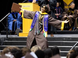 Student wearing Adelphi graduation attire with his arms out and smiling on stage at the Commencement ceremonies. 