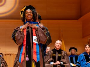 A student in cap and gown smiles broadly with her hands on her chest as she receives the hood awarded PhD recipients.