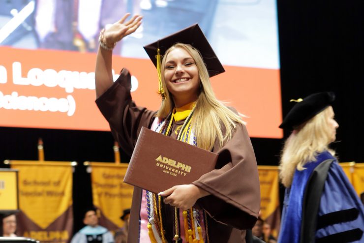 Individual waving at graduation ceremony wearing brown gown with gold accessories.