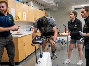 A professor and two graduate exercise science students observe a client on a stationary bicycle, and they seem to be encouraging him to keep pedaling.