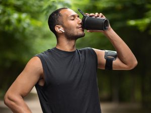 A man in workout gear pauses on a tree-lined path to drink from a shaker bottle, wearing an armband phone holder and wireless earbuds.