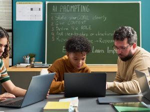 A teacher helps two students on laptops in a classroom with a chalkboard listing steps for AI prompting: state your goal, give context, define the role, ask step-by-step, and revise.