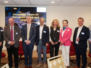 A group of eight professionals in business attire pose together in a conference room in front of a screen displaying "Leadership Forum on Successful Aging and Longevity."