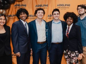  Six students of different genders and ethnicities smile facing the camera. There are brown and gold balloons on the left and they stand in front of a gold background with the words “Adelphi University.”