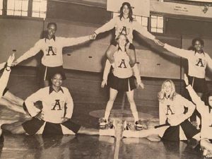 Adelphi cheerleaders in formation on the basketball court. Four cheerleaders are seated on the floor, five are behind them, with one cheerleader carrying another on her shoulders.