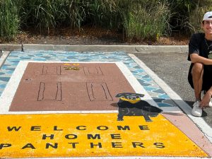 A young man, James Tullo, kneels by his parking spot design, labeled "Welcome Home Panthers"