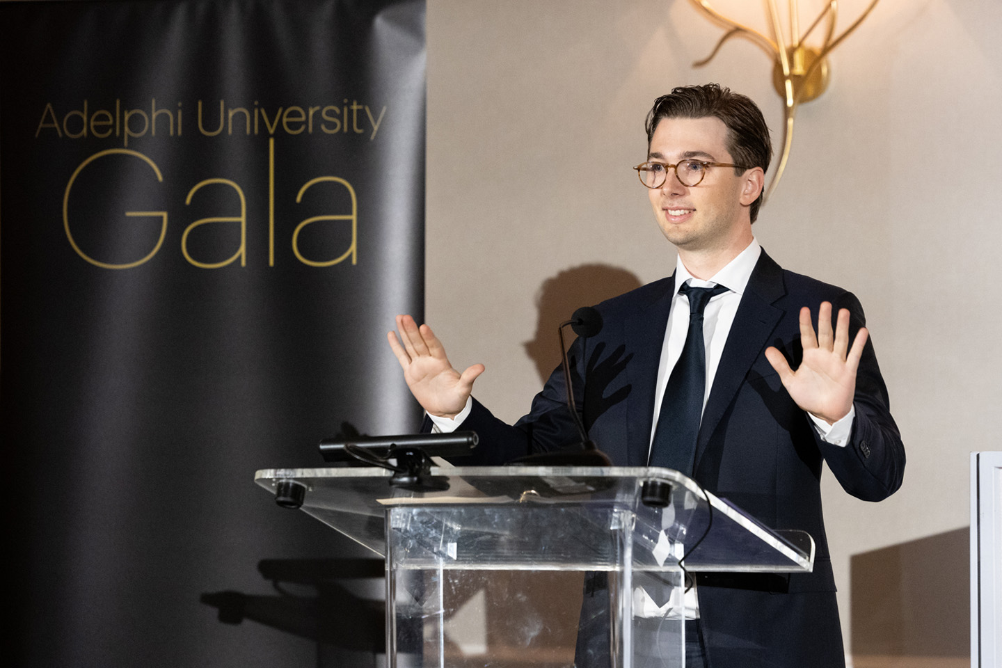 A formally dressed young man stands at the podium, gesturing with both hands.
